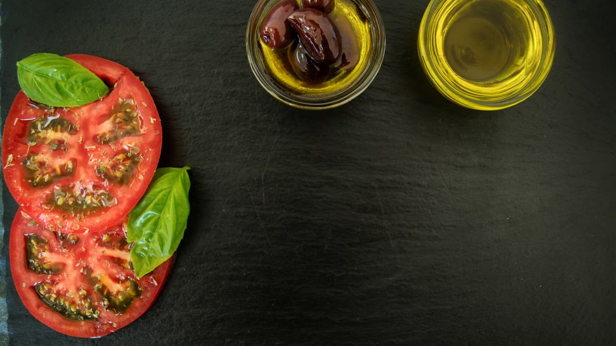 Top view of fresh basil, sliced tomatoes, olives, and olive oil on slate, perfect for Italian cuisine themes.